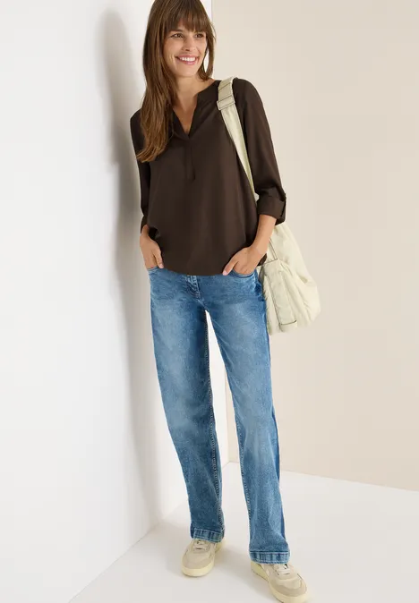 Frau mit brauner Bluse, Jeans und beiger Tasche lächelt.